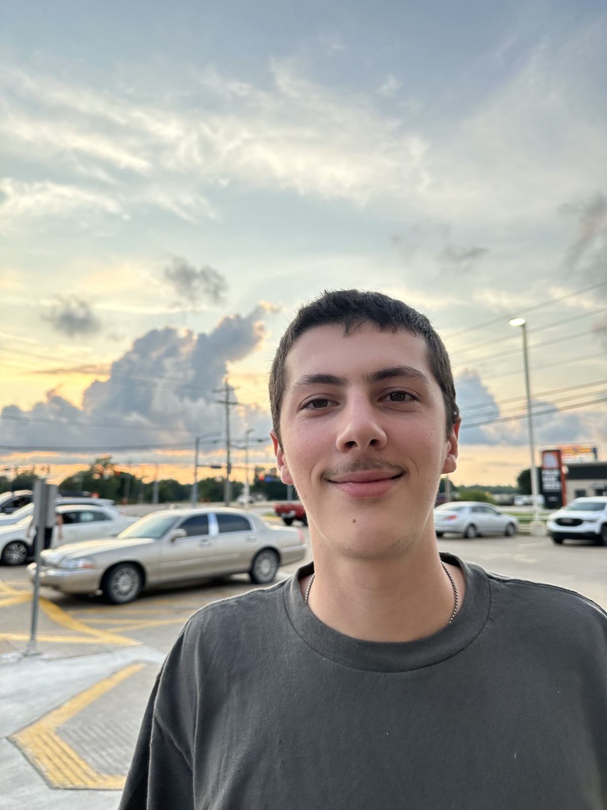 Charlie smiling outdoors at sunset, classic cars and a dramatic cloud-filled sky behind him