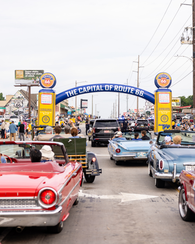 Vertical shot of classic cars driving under the Capital of Route 66 arch, spectators lining the street