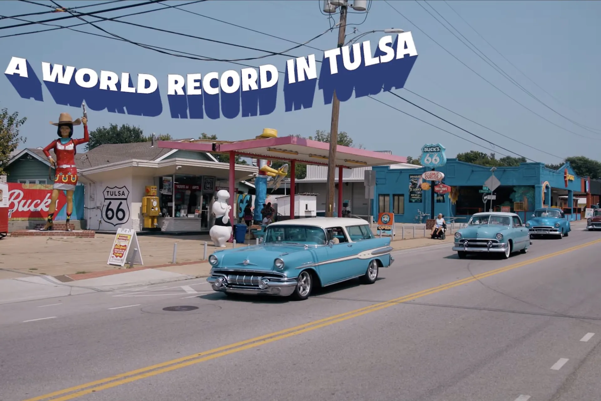 A classic light-blue wagon and other vintage cars driving down historic Route 66 in Tulsa, past retro roadside attractions, with 'A World Record in Tulsa' captioned above