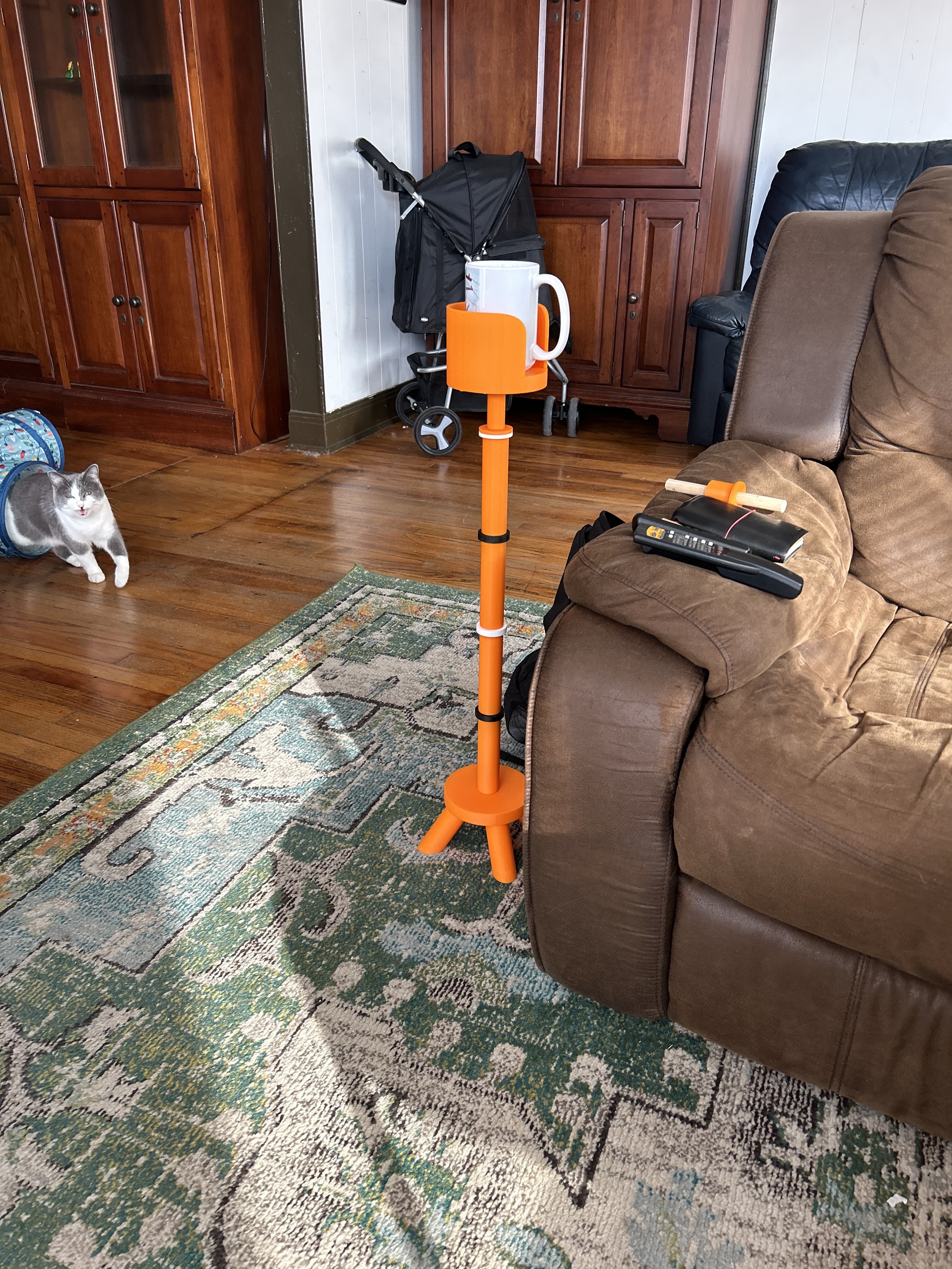Orange 3D-printed tripod stand holding a mug at couch-arm height, positioned next to the recliner